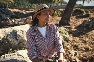 Kareli gömlek ve hasır şapka giymiş kayalık arazide oturan bir kadın, sakin bir açık hava ortamının tadını çıkarıyor. Doğal güzellik ve rahatlama, güneş ışığı ve doğa tarafından benimsenen anahtar temalardır..