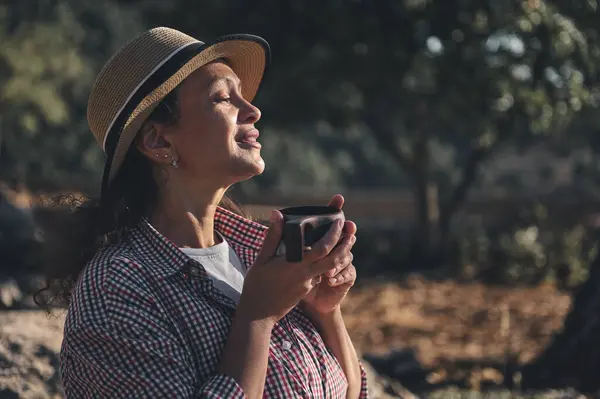 Hasır şapkalı bir kadın, elinde bir fincan kahveyle sakin bir sabah geçirir. Huzurlu ifade, doğanın ortasında rahatlamayı ve memnuniyeti, sükuneti kucaklamayı önerir..