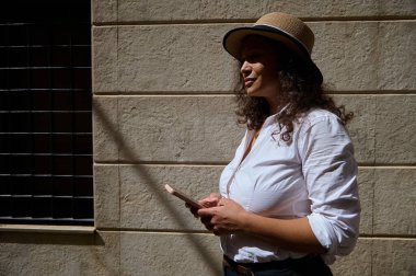 Hasır şapkalı ve beyaz tişörtlü bir kadın güneşli bir şehir duvarına karşı duruyor, akıllı bir telefonla düşünceli bir şekilde..