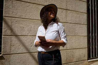 Bir kadın elinde akıllı telefonuyla güneşli bir duvara dayanıyor. Şapka takıyor ve rahat görünüyor, güneşli günün tadını çıkarıyor. Günlük kıyafetleri ve ifadesi boş zaman ve bağlantı olduğunu gösteriyor..