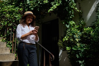 Gündelik giysiler içindeki bir kadın, açık havada güneşli bir günün tadını çıkarır, akıllı telefonunu yemyeşil bir merdivenin yanında kullanır. Gülüşü ve gevşemiş tavırları bir eğlence ve bağlantı hissi veriyor..