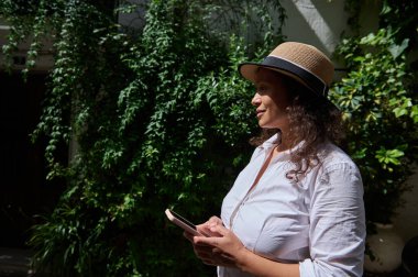 Hasır şapkalı ve beyaz gömlekli bir kadın, elinde tabletle güneşli bir günün tadını çıkarıyor. Yeşillik ve doğal ışık huzurlu ve huzurlu bir atmosfer yaratır..