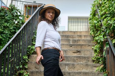 Şık bir şapka giyen kendine güvenen bir kadın, yemyeşil bir bahçenin merdivenlerinde duruyor. Onun rahat duruşu ve anlamlı bakışları kendine güven ve sükunet hissi veriyor..