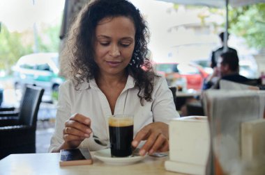 Beyaz gömlekli bir kadın açık bir kafede oturup bir fincan kahvenin tadını çıkarıyor. Rahatlamış ve mutlu görünüyor, huzurlu bir anın tadını çıkarıyor. Güneşli hava ve şehir hayatı etrafını sarmış..