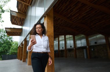 Beyaz gömlekli şık bir kadın ahşap bir mimari yapının altında dururken akıllı telefon kullanıyor. Açık hava teknolojisi ve modern tasarım..