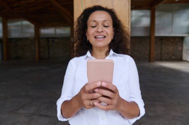 A woman wearing a white shirt is smiling as she uses her smartphone in an indoor setting. The rustic indoor environment adds a cozy feel, conveying relaxation and connectivity. clipart