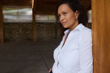 A contemplative woman in a white shirt leans against a wooden post in an indoor setting. Her thoughtful expression suggests introspection. The natural background adds warmth to the scene. clipart