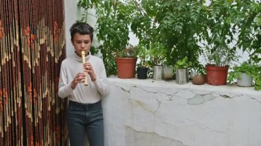 Genç bir çocuk kapalı bir alanda flüt çalarak duruyor. Arka planda yemyeşil bitkiler ve dekoratif boncuklu bir perde yer alıyor. Huzurlu bir atmosfer yaratıyor..