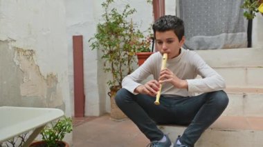 Genç bir çocuk açık havada bir basamağa oturmuş, odaklanmış bir flüt çalıyor. Kırsal çevre, sakinlik ve yaratıcılık hissi uyandırıyor..
