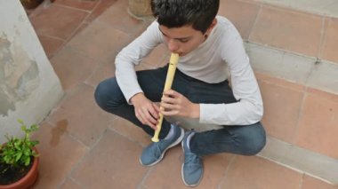 Terasta oturan genç bir çocuk, sarı kayıt cihazını çalmaya odaklanmış. Ortalık sakin ve açık hava müziğinin bir göstergesi olan saksılarla dolu..