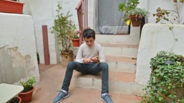 Sıradan giyinmiş genç bir çocuk, akıllı telefonuna odaklanmış, açık hava basamaklarında oturuyor. Etrafı çeşitli saksı bitkileriyle çevrili, sakin ve sakin bir avluya gömülmüş görünüyor..