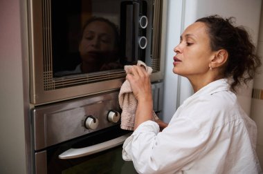 Bir kadın özenle fırını temizler, odak noktası gösterir ve ev işlerinde özen gösterir. Detaylara olan ilgisi, modern mutfak ortamındaki temizlik ve adanmışlığı vurguluyor..