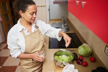 A woman in an apron prepares a healthy meal in her kitchen, adding ingredients to a dish. Fresh vegetables are visible on the counter, creating a cozy and wholesome atmosphere. clipart