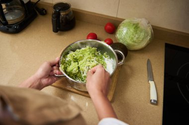 Eller, mutfak ortamında marul, avokado ve domates gibi malzemelerin yanı sıra mutfak aletleri içeren lezzetli taze marul salatası hazırlıyor..
