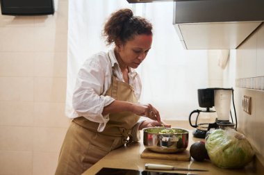 Önlük takan bir kadın paslanmaz çelik bir kasede taze malzemeler karıştırıyor. Parlak bir mutfakta salata yapıyor. Sağlığı, ev yemeklerini ve aşçılık tutkusunu somutlaştırıyor..