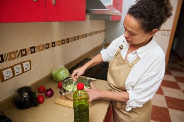Bir kadın taze salata hazırlarken mutfakta duruyor. Etrafı sebzelerle çevrili malzemeleri özenle karıştırmaya odaklanmış. Mutfak aydınlık ve düzenli..