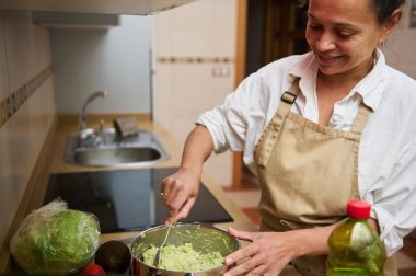 Mutfakta guacamole yaparken gülümseyen bir kadın. İçindekiler avokado ve sebzeler. Mutfak yaratıcılığı ve sağlıklı beslenme konsepti.