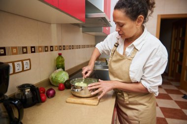 Önlüklü bir kadın, sebzeleri tencerede karıştırıyor ve modern mutfak ortamında taze bir salata hazırlıyor..