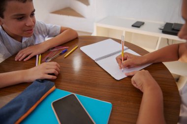 İki çocuk bir çalışma oturumunda bir masada oturuyorlar. Biri bir deftere yazarken diğeri dikkatlice izliyor. Kalemler ve okul malzemeleri masaya saçıldı..