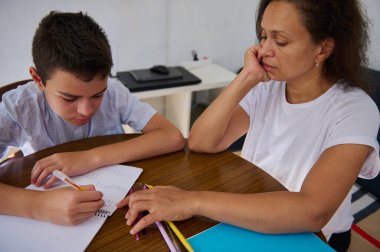 Bir anne tahtadan bir masada oğluna ev ödevinde yardım eder. Çocuk, annesi onu desteklerken matematik problemlerini deftere yazmaya odaklanmış..
