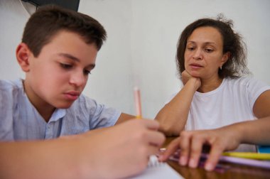 Bir anne, oğlunun mutfak masasında ödevine özenle yardım eder. Anne ona rehberlik ederken çocuk yazmaya odaklanıyor, evde bir öğrenme ve destek sahnesi sergiliyor..