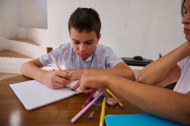 Genç bir çocuk, ahşap bir masanın üzerindeki renkli kırtasiye malzemeleriyle çevrili bir yetişkin ona rehberlik ederken ev ödevine yoğun bir şekilde odaklanır..