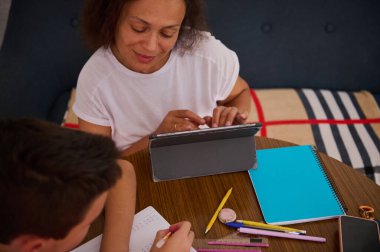 Bir kadın ve çocuk, ahşap bir masanın üzerinde tablet, not defteri ve kırtasiye ile öğrenim görerek rahat bir ortamda ortak bir eğitim deneyimini teşvik ediyorlar..
