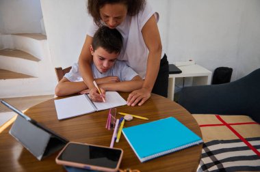 Bir anne sevgiyle oğluna tahtadan bir masada ödevlerinde yardım eder, öğrenme ve destek olur. Tabletler ve defterler dağınık, rahat bir çalışma ortamı yaratıyorlar..