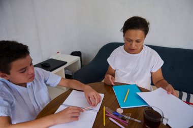 Bir anne ve oğlu tahtadan bir masada oturuyorlar, evde eğitim görüyorlar. Not defterleri, kalemler ve bir fincan çay odaklanmış ve eğitici bir atmosfer izlenimi veriyor..