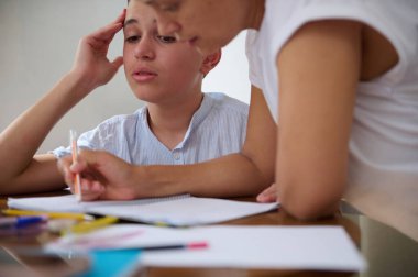 Bir anne mutfak masasında oğluna ev ödevinde yardım eder. Çocuk görevini tamamlarken odaklanmış görünüyor. Kalemler ve kağıtlar masanın etrafına saçılmış..