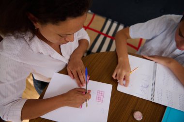 Bir anne çocuğuna ahşap bir masada matematik ödevinde yardım eder, sıcak bir ev ortamında öğrenme ve destek sağlar, aile eğitimini ve birlikteliği betimler..