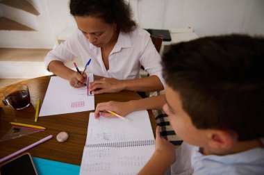 Bir anne ve oğul mutfak masasında matematik ödevlerinde işbirliği yaparak öğrenme ve iletişimi teşvik ediyorlar. Kalemler, kağıtlar ve odak noktaları açık, eğitimi ve aile bağlarını ön plana çıkarıyor.