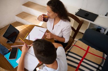 Bir anne çocuğuna ev ödevlerinde yardım eder, öğrenme ve aile bağlarını teşvik eder. Not defterleri ve tabletlerle dolu bir masada oturup çay içip ders çalışırlar..
