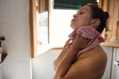 Bir kadın sabah cilt bakımına alışır, yumuşak bir havluyla boynunu okşar. Pencereden güneş ışığı süzülüyor ve güne huzur dolu ve ferahlatıcı bir başlangıç yaptığını vurguluyor..