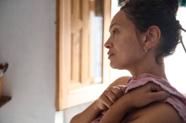 Havluya sarılı sakin bir kadın düşünceli bir şekilde bakar, evde sakin bir sabahın tadını çıkarır. Doğal ışık onun barışçıl ifadesini vurgular ve kırsal iç kısımları sahneye sıcaklık katar..