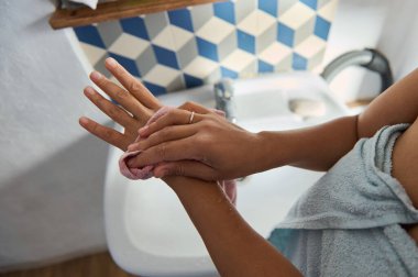 Bir kadın ellerini nazikçe lavabonun üzerinde havluyla kurutuyor. Fayanslı banyo arka planı estetik bir dokunuş katıyor. Sahne temizliği ve kişisel bakımı her gün sunuyor..