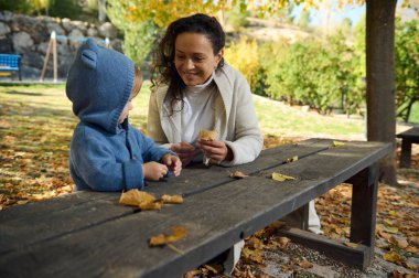 Genç bir anne ve çocuğu, güneşli bir sonbahar gününde parkın yaprakları üzerinde neşe ve doğayı kucaklayarak birbirlerine bağlanırlar..