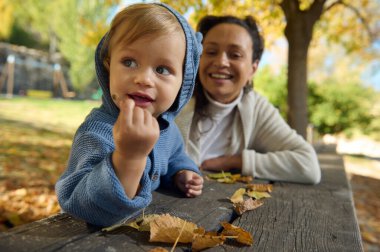 Mavi kazak giyen neşeli bir çocuk, gülen bir yetişkinin eşlik ettiği bir park masasında sonbahar yapraklarının keyfini çıkarıyor. Sahne sıcaklığı, birlikteliği ve açık havada düşüşün güzelliğini yansıtıyor..