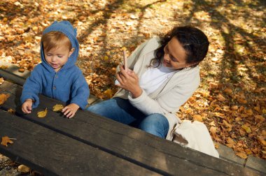 Bir anne ve çocuğu, sonbahar yapraklarıyla çevrili bir piknik masasında neşeli bir anı paylaşıyorlar. Bebek düşen yaprakları merak ederken anne de sıcaklığı ve bağlantıyı somutlaştıran telefonuyla sahneyi yakalıyor..