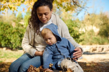 Sevgi dolu bir anne, güneşli bir sonbahar gününde bebeğiyle oturur. Etrafı yapraklarla çevrilidir. Sıcak atmosfer, açık havada bir şefkat ve bağ anı yakalar..