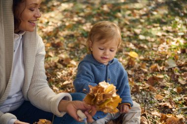 Neşeli bir anne ve küçük çocuğu güneşli bir parkta sonbahar yapraklarıyla oynuyorlar, neşeli bir anı paylaşıyorlar..