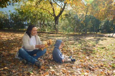 Bir anne ve çocuğu, renkli sonbahar yapraklarıyla çevrili, güneşli bir günün tadını çıkarırlar. Görüntü, neşe verici bir bağ, kahkaha ve doğanın güzelliğini yakalar..