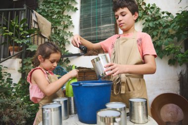 Yaratıcı bir bahçe işiyle uğraşan iki çocuk teneke kutuları toprakla dolduruyor. Açık alandalar, yemyeşil bitkilerle çevrili, eğlenceli, aktif bir projede işbirliği yapıyorlar..