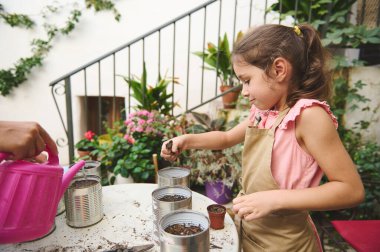 Genç bir kız, yeşillik ve çiçeklerle çevrili güneşli bir avluda renkli kutulara tohum ekiyor. Bahçıvanlığa odaklanması yaratıcılığı, büyümeyi ve doğayla bağ kurmanın neşesini yakalıyor..