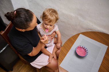 Bir anne, bebeğini sevgiyle mutfakta besliyor. Çocuk annesinin kucağında otururken bir kaşık dolusu sağlıklı pembe smoothie 'nin tadını çıkarıyor. Sıcak bir aile anı..