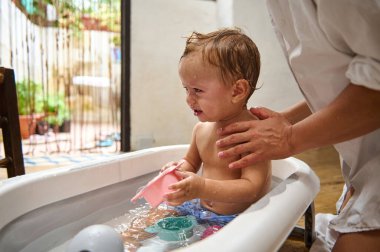 Küçük bir çocuk, sıcak, sıcak bir banyoda ebeveynleri tarafından desteklenirken banyo sırasında ağlar. Bebek, su ve banyo oyuncaklarıyla çevrili beyaz bir küvette otururken pembe bir bardak tutuyor..
