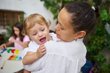 Sevgi dolu bir anne neşeli bebeğini tutarken başka bir çocuk bahçede renkli oyuncaklarla oynuyor..
