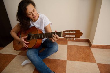 Bir kadın damalı zeminde oturmuş, odaklanarak akustik gitar çalıyor. Sahne, müziğe duyulan rahatlama ve tutkuyu aktarıyor. Kişisel zevkin sakin bir anını vurguluyor..