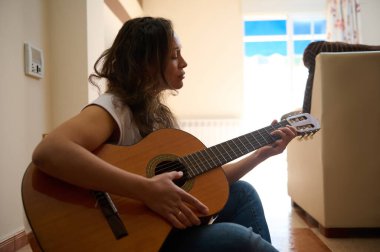 Bir kadın rahat bir ev ortamında yerde otururken akustik gitar çalmaktan hoşlanır. Doğal ışık, müziğine odaklanırken sıcak bir atmosfer yaratır..