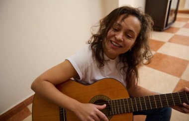 Bir kadın fayanslı zeminde oturmuş, neşeli bir şekilde akustik gitar tıngırdatıyor. Onun rahat duruşu ve parlak gülüşü mutluluk ve müzikal tutkuyu ifade eder..